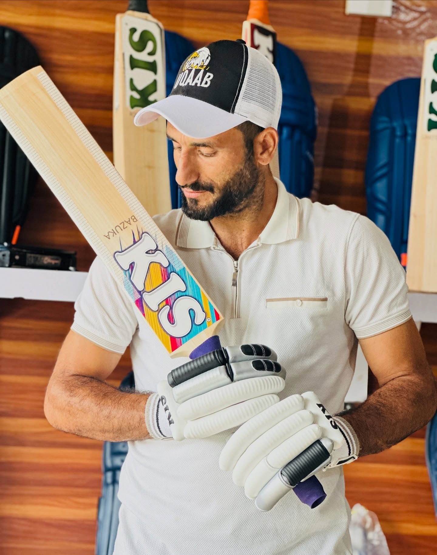A man in a white polo shirt and a black-and-white cap holds a wooden cricket bat with a colorful graphic label. He is wearing white cricket gloves in what appears to be a shop or studio setting with wooden walls and sports gear in the background.
