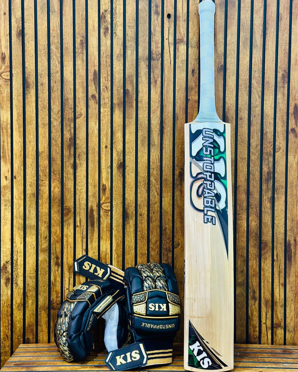 A cricket bat and matching cricket gloves displayed against a vertical wooden slat wall. The bat is light natural wood with blue, white, and black graphics reading 'UNSTOPPABLE', and the gloves are dark navy with gold and white 'KIS' and 'UNSTOPPABLE' branding.