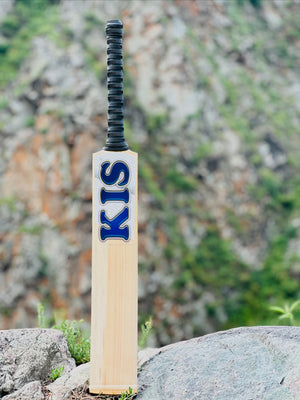 A Kashmir willow cricket bat standing upright on rocks outdoors, with a blurred rocky hillside in the background. The bat is light natural wood with a black handle grip and blue 'KIS' lettering outlined in silver on the blade.