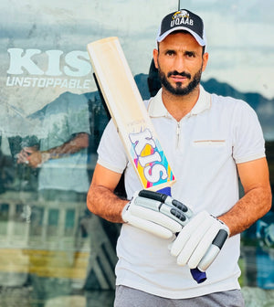 A man wearing a white polo shirt, white cricket gloves, and a black cap with a gold logo holds a wooden cricket bat with colorful 'KIS' branding Bazuka Edition. The background is a reflective glass wall showing a blurred version of the same person and the text 'KIS UNSTOPPABLE'.