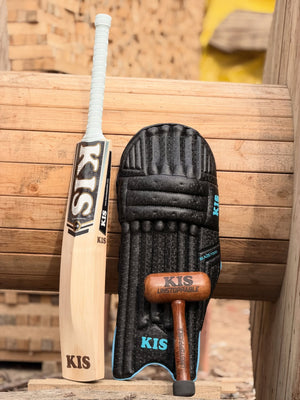 A KIS brand cricket bat and a black KIS batting pad with a wooden mallet, displayed together on a rustic wooden bench with a blurred brick wall background.