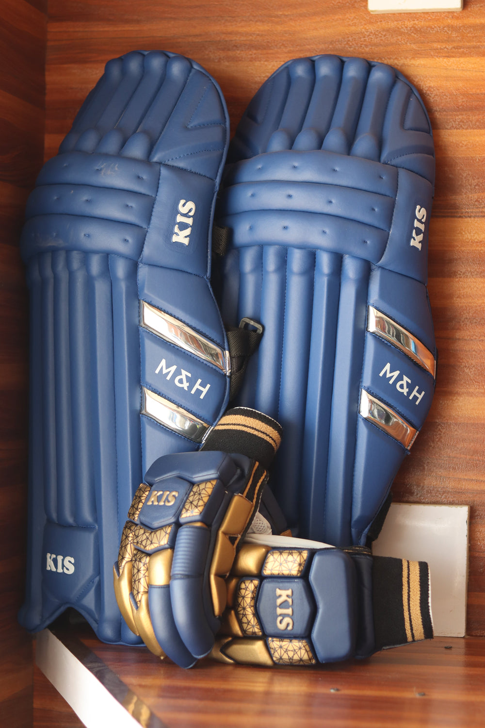A pair of blue cricket pads with gold and black accents, including a matching wrist guard, displayed on a wooden surface. The brand names 'KIS' and 'M&H' are clearly visible on the gear.